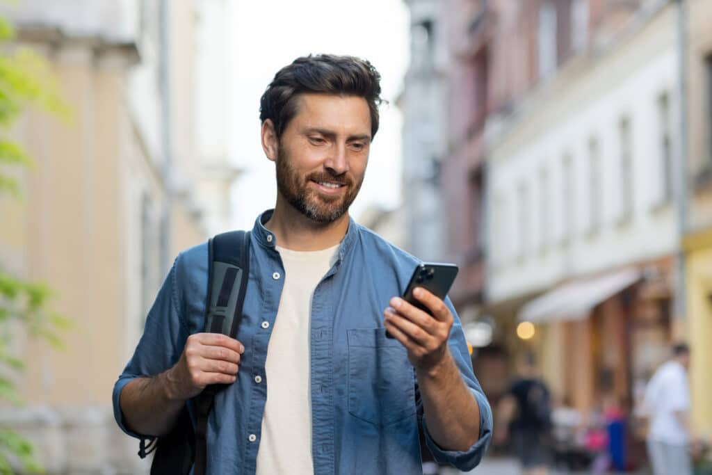 A handsome smiling young man wearing a denim shirt and carrying a backpack is standing on a city street and using a mobile phone A handsome smiling young man wearing a denim shirt and carrying a backpack is standing on a city street and using a mobile phone