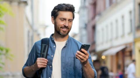 A handsome smiling young man wearing a denim shirt and carrying a backpack is standing on a city street and using a mobile phone A handsome smiling young man wearing a denim shirt and carrying a backpack is standing on a city street and using a mobile phone